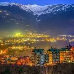 Manali town at night with snow-covered Himalayan peaks in the background.